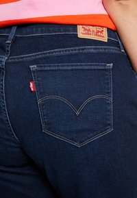 Jeans de mezclilla azul oscuro que presentan un bolsillo trasero característico con un diseño de costura en forma de corazón y una etiqueta de cuero marrón con la marca.