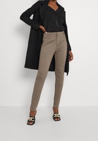 Bruin geruite broek met een getailleerde pasvorm, gecombineerd met een zwart satijnen top en een lange zwarte jas. Zwarte sandalen met hoge hakken hebben gouden accenten.