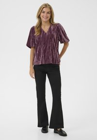 Violette, kurzärmlige V-Ausschnitt-Bluse mit einem strukturierten, gestreiften Muster, kombiniert mit schwarzen Schlaghosen und schwarzen Loafers.