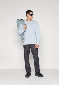 Sweater bleu deschis, pantaloni negri și pantofi negri. Un rucsac gri este aruncat pe o umăr. Ochelari de soare aviator cu lentile albastre.