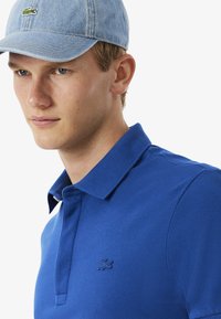 Kungligt blå pikétröja i texturerat tyg med krage, med en subtil, broderad logotyp på bröstet. Matchas med en ljusblå keps.