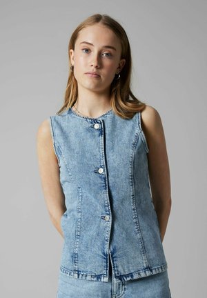 Noor naine õlgadeni juustega, kes kannab helesinist, varrukateta, nööbitavat teksavesti ja sobivaid teksapükse, seisab halli tausta ees.