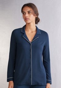 Mörkblå långärmad pyjamasöverdel med vit piping, knäppning fram och krage. Mjukt material med en slät yta.