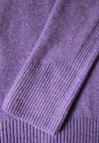 Detail eines lila Wollpullovers mit geripptem Saum und Bündchen, weicher Textur und warmem, leicht meliertem Erscheinungsbild.