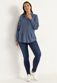 Denim chambray shirt with vertical pleats, button-down front, long sleeves, semi-fitted waist, and a curved hem. Worn with dark jeans and sneakers.