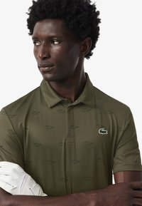 Polo verde oliva con un sutil patrón de cocodrilos en toda la prenda, con cuello y un pequeño logo bordado en el pecho. Textura suave.
