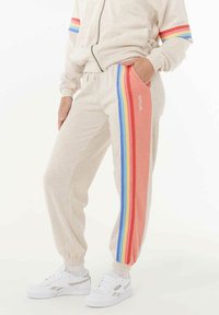 Persoon in beige sweatpants met meerkleurige regenboogstrepen aan de zijkant en witte sneakers, met een hand in de zak.