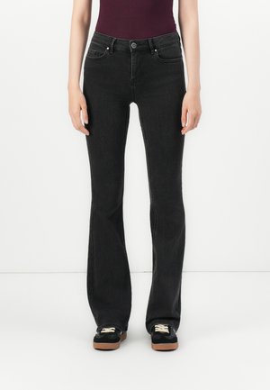 Γυναίκα που φοράει μαύρα τζιν με ψηλή μέση και καμπάνα και μαύρα αθλητικά παπούτσια με λευκά κορδόνια, στέκεται μπροστά σε λευκό φόντο.