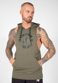 Olivgrüner ärmelloser Hoodie mit Fronttasche, ausgestattet mit einem kreisförmigen Grafik-Logo. Schwarze Kordeln und strukturierte Stoffdetails.