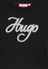 Felpa nera con un grande logo argento "Hugo" in carattere corsivo. Tessuto a coste sul colletto e sull'orlo, con dettaglio della tag rossa all'interno.