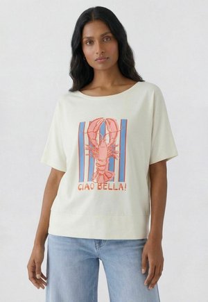 Frau trägt ein cremefarbenes T-Shirt mit einem roten Hummer und blauen vertikalen Streifen, beschriftet mit "CIAO BELLA!", kombiniert mit hellblauen Jeans.