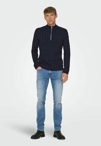 Marineblaue Strickpullover mit hohem Kragen und halbem Reißverschluss, kombiniert mit hellblauen Jeans und schwarzen Stiefeln. Glatte Textur und taillierte Passform.