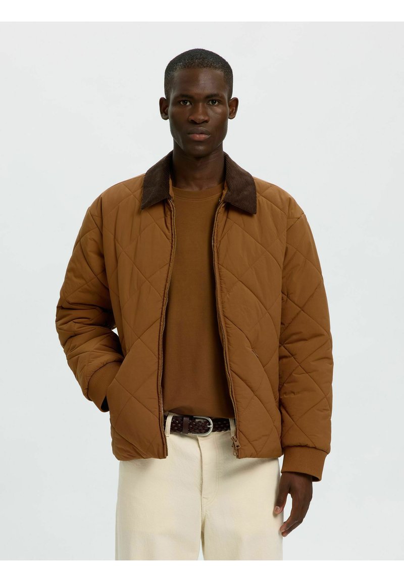 Jeune homme portant une veste matelassée marron avec un col foncé, une chemise marron, un pantalon beige clair et une ceinture tressée foncée, debout devant un fond uni.