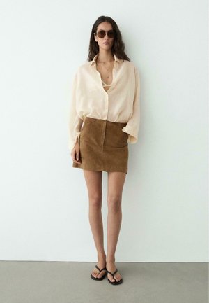 Kvinde iført oversized cremefarvet bluse, brun ruskind mininederdel, sorte flipflops, solbriller, står op ad en hvid væg.