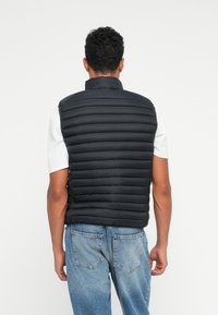 Sort polstret vest med høj krave, der har et horisontalt quilted design og er ærmeløs, parret med en hvid skjorte og blå jeans.