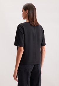 Top nero a maniche corte con scollo quadrato, tessuto liscio e taglio dritto. Il retro presenta una cucitura per una vestibilità strutturata.