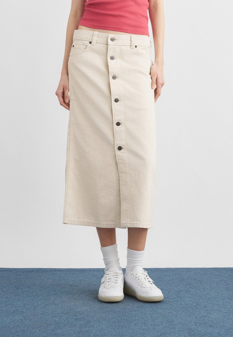 Femeie purtând o fustă midi bej cu nasturi în față, adidași albi și șosete albe, stând pe o mochetă albastră pe un fundal simplu alb.