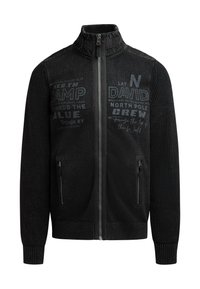 Schwarze Jacke mit Reißverschluss und hohem Kragen, strukturierten Ärmeln, Fronttaschen und bedruckten Grafiken in verschiedenen Schwarz- und Grautönen.