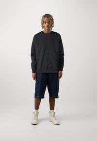 Mladý člověk s blond dreadlocky, oblečený v černé mikině, tmavých denimových šortkách, bílých ponožkách a krémových vysokých teniskách, stojící čelem.