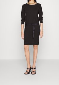 Schwarzes Kleid mit langen Ärmeln und taillierter Form, versehen mit einem Gürtel mit Logodetails. Kombiniert mit schwarz-geschlossenen High Heels mit Riemchen.