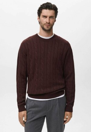 Suéter de punto trenzado burdeos con cuello redondo, puños y dobladillo de canalé; superpuesto a una camiseta blanca y acompañado de pantalones grises.