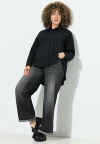 Zwart overhemd met een gerimpelde zoom, gecombineerd met donkergrijze jeans met studaccenten. Model draagt zwarte instappers.