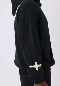 Sudadera negra con capucha, con tela desgastada y un parche de estrella blanca en la manga derecha. Textura suave y diseño de corte relajado.