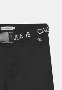 Černé džíny s pevným materiálem, výrazným páskem s nápisem "CALVIN KLEIN JEANS" vytištěným bílou barvou a stříbrným logem "ck" na přední straně.