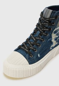 Aukštai cortezi stiliaus sportbačiai iš nusidėvėjusio mėlyno džinsinio audinio su juodomis raišteliais ir Marc Jacobs prekės ženklu. Balta guma puošia pirštų apsaugą ir tekstūruotą padas.