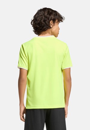 Junge Person mit lockigem Haar, die ein leuchtend gelbes kurzärmeliges Shirt und schwarze Shorts trägt, mit dem Rücken zum Betrachter vor einem schlichten weißen Hintergrund.