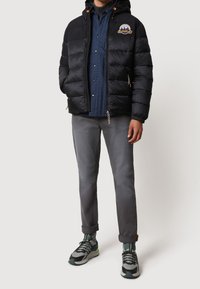 Sort pufferjakke med hætte og lynlåslommer, båret over en blå ternet skjorte, parret med grå bukser og grå-grønne sneakers.