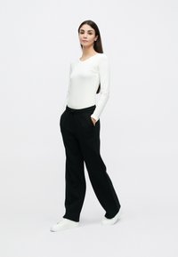 Λευκή μπλούζα με μακριά μανίκια συνδυασμένη με μαύρο, χαλαρό παντελόνι με ελαστική ζώνη μέσης και τσέπες, συμπληρωμένο με λευκά αθλητικά παπούτσια.