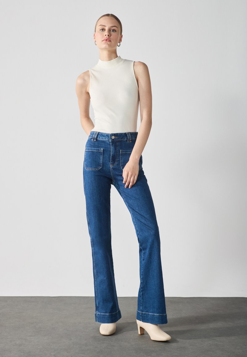 Gerippetes weißes ärmelloses Oberteil kombiniert mit hochgeschnittenen blauen Schlagjeans, die Rückentaschen und eine helle Waschung aufweisen, abgerundet mit cremefarbenen High Heels.