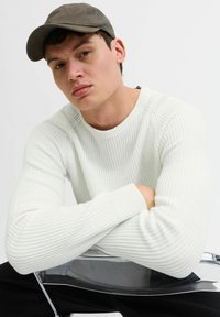 Maglione bianco a coste con scollo rotondo, abbinato a un cappello scuro. Braccia incrociate, seduto su una sedia trasparente contro uno sfondo neutro.