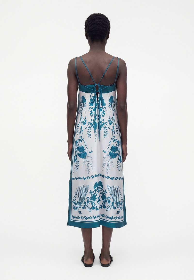 Woman wearing a sleeveless white midi dress with teal floral patterns and thin straps tied at the back, standing against a plain background.