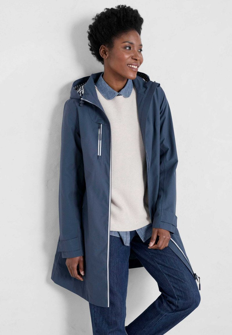 Granatowy wodoodporny płaszcz z zamkiem błyskawicznym z przodu i kapturem, z kontrastowymi szwami, zamykanymi kieszeniami i luźnym krojem na beżowy sweter.