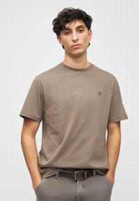 Brązowy bawełniany t-shirt z okrągłym dekoltem, krótkimi rękawami i małym wyszywanym detalem na piersi. Noszony z szarymi spodniami i paskiem.