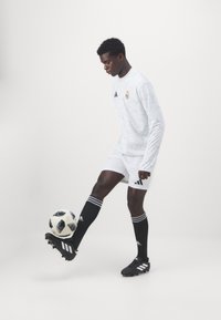 Hvid langærmet trøje med et subtilt mønster, sorte shorts og sorte strømper med hvide striber; spilleren balancerer en sort og hvid fodbold.