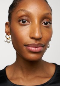 Close-up of a face with smooth skin, brown eyes, and natural makeup, wearing gold hoop earrings with three white pearl accents.