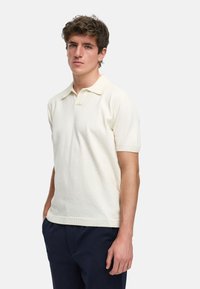 Kortermet polo skjorte i off-white, laget av teksturert stoff. Har klassisk krage og ribbestrikket kant. Båret med marineblå bukser.