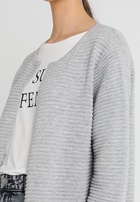 Světle šedý pletený kardigan s žebrovanou texturou, otevřený vpředu bez zapínání, vrstvený přes bílé tričko s černým textem.