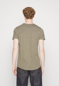 Muž s kudrnatými červenými vlasy, oblečený v těsném olivově zeleném tričku a tmavě modrých džínách, stojící čelem k čisté bílé zdi.