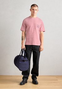 Stripete rød og hvit T-skjorte med brystlomme, parret med mørke denimjeans. Holder en rund marineblå duffelbag med doble håndtak.