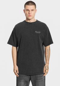 Czarny oversize'owy t-shirt z okrągłym dekoltem. Posiada wyblakłą fakturę i mały biały nadruk z przodu. Noszony z szarymi dżinsami.