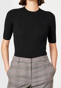 Musta ribbineulottu lyhythihainen paita pyöreällä kaula-aukkolla, yhdistetty korkeavyötäröisiin ruutukuosisiin housuihin, joissa on vaaleanpunaisia ja harmaita yksityiskohtia.