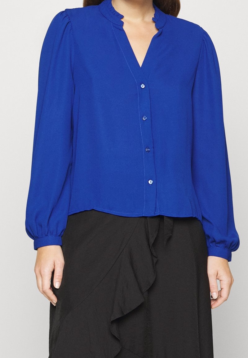 Blusa blu con maniche lunghe a sbuffo, colletto ruffled e chiusura con bottoni. Il tessuto appare liscio con una finitura leggermente lucida.