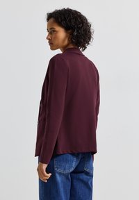 Blazer bordeaux realizzato in tessuto morbido con colletto e due bottoni, abbinato a jeans in denim blu. Dettagli cuciti visibili sulle tasche.