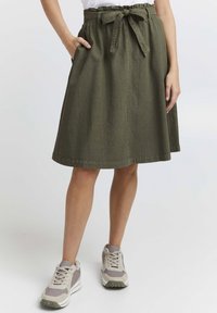 Olive green, knee-length skirt made of textured fabric, featuring a waistband with a fabric tie and side pockets. Styled with athletic sneakers.