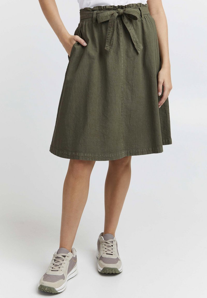 Olive green, knee-length skirt made of textured fabric, featuring a waistband with a fabric tie and side pockets. Styled with athletic sneakers.