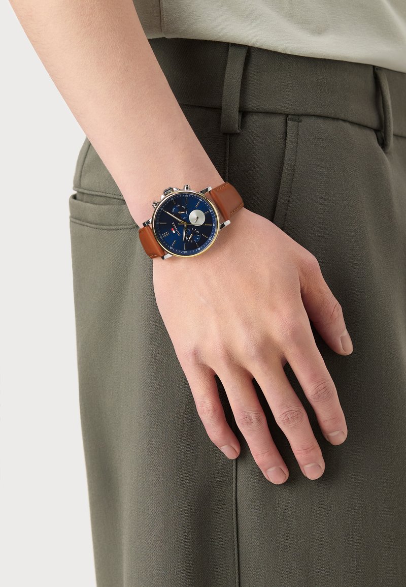 Montre avec bracelet en cuir marron, cadran bleu, boîtier argenté et fonctions chronographe. La montre est portée sur une main posée sur un pantalon vert.